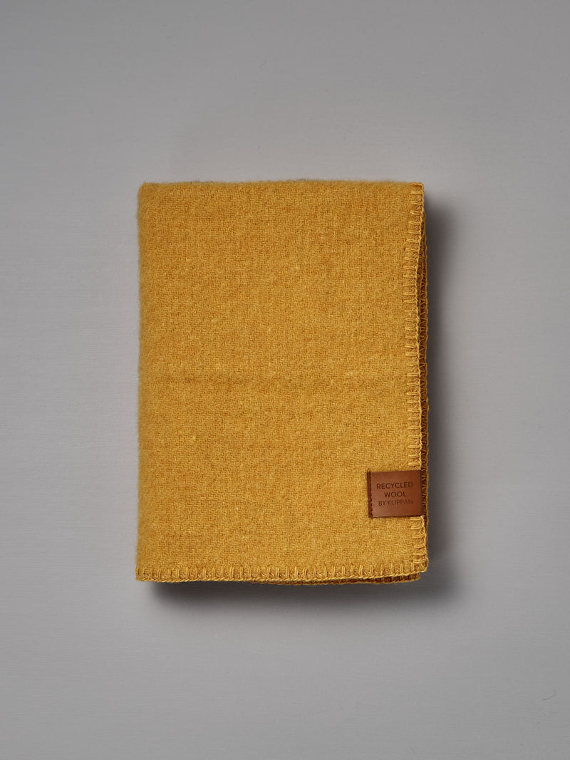 Folded True Baby Blanket – Straw, made from yellow recycled wool and featuring a brown “Recycled Wool by Re-born” label, crafted by Klippan Sweden, placed on a plain grey surface.