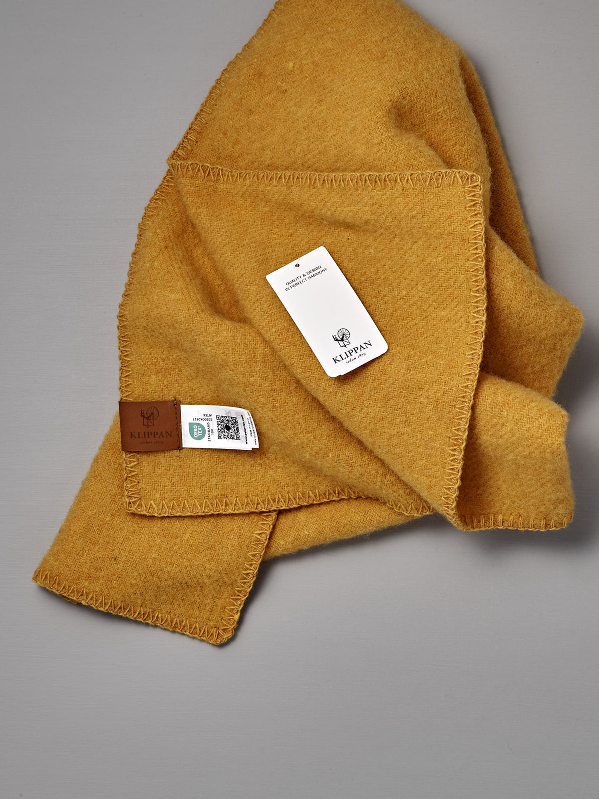 A folded yellow wool blanket, the True Baby Blanket in Straw, with a stitched edge, product tags, and a leather Klippan Sweden label is shown on a gray background. Crafted with the same attention to quality as Klippan’s renowned recycled wool blankets and eco lambswool textiles, this baby blanket exemplifies timeless Scandinavian design.