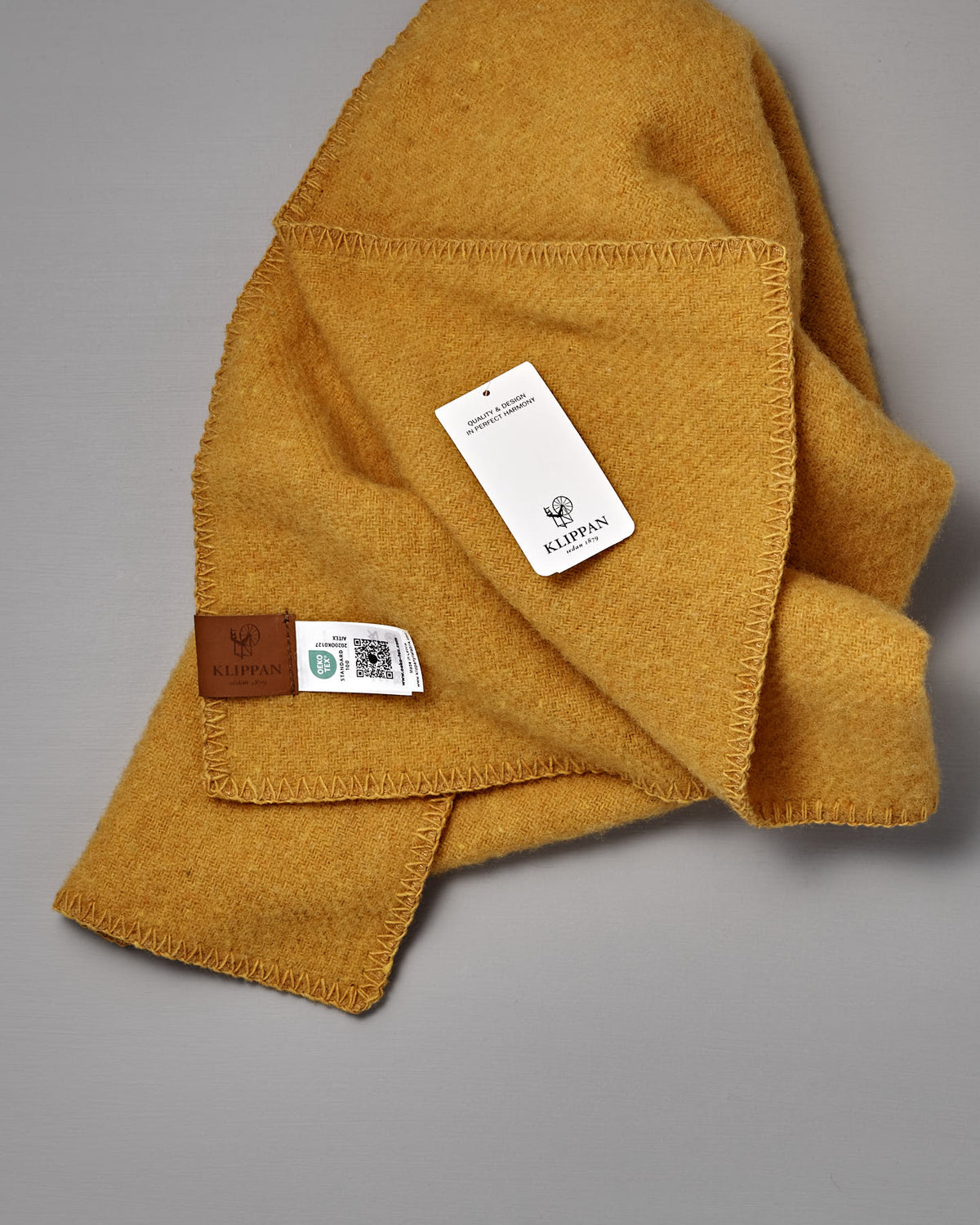 A folded yellow wool blanket, the True Baby Blanket in Straw, with a stitched edge, product tags, and a leather Klippan Sweden label is shown on a gray background. Crafted with the same attention to quality as Klippan’s renowned recycled wool blankets and eco lambswool textiles, this baby blanket exemplifies timeless Scandinavian design.