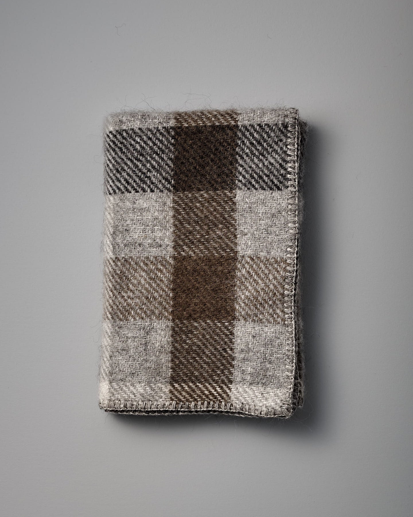 A Gotland Wool Baby Throw – Multi Grey, featuring a classic brown, gray, and beige plaid pattern inspired by timeless Klippan of Sweden designs, is displayed on a plain gray surface.