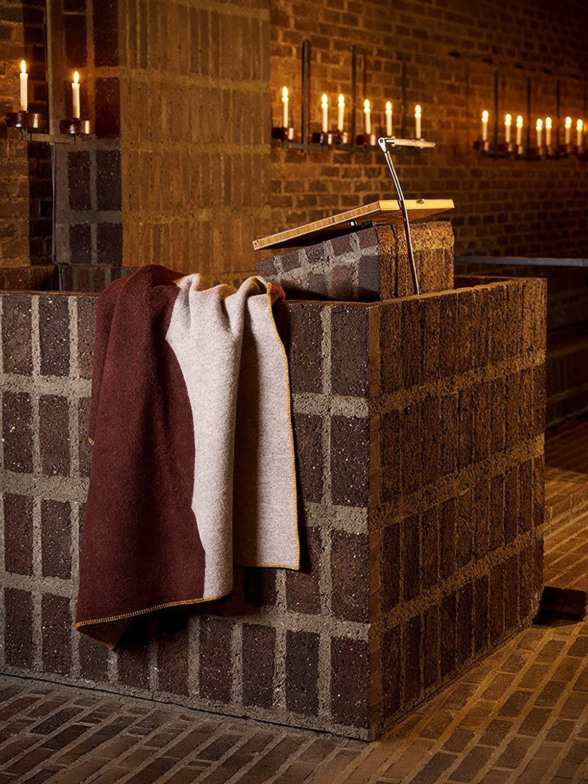 A Sankt Petri Facade Wool Throw – Dark Brick is draped over a brick pulpit with a bookstand in a dimly lit church. This cozy organic wool throw, inspired by Kristina Stark’s design, adds warmth as candles line the brick wall in the background.
