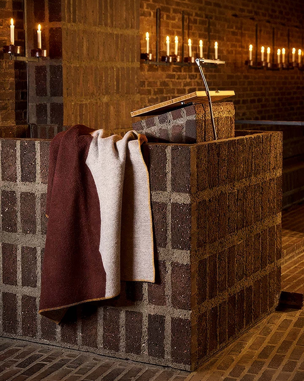 A Sankt Petri Facade Wool Throw – Dark Brick is draped over a brick pulpit with a bookstand in a dimly lit church. This cozy organic wool throw, inspired by Kristina Stark’s design, adds warmth as candles line the brick wall in the background.