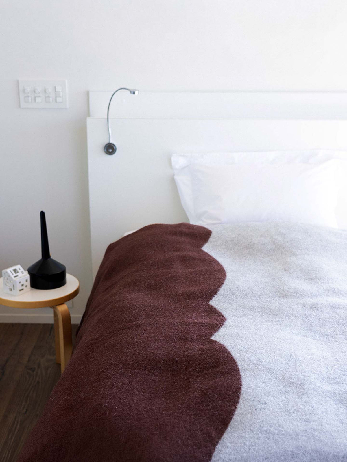 A neatly made bed features a Sankt Petri Facade Wool Throw in Dark Brick, complemented by a white pillow and a headboard with a reading light. A small wooden side table holds a black object and two small white items, adding to the room’s simple elegance.