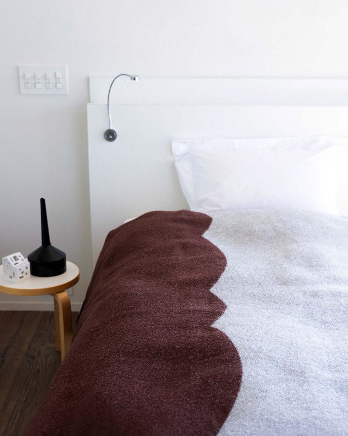 A neatly made bed features a Sankt Petri Facade Wool Throw in Dark Brick, complemented by a white pillow and a headboard with a reading light. A small wooden side table holds a black object and two small white items, adding to the room’s simple elegance.