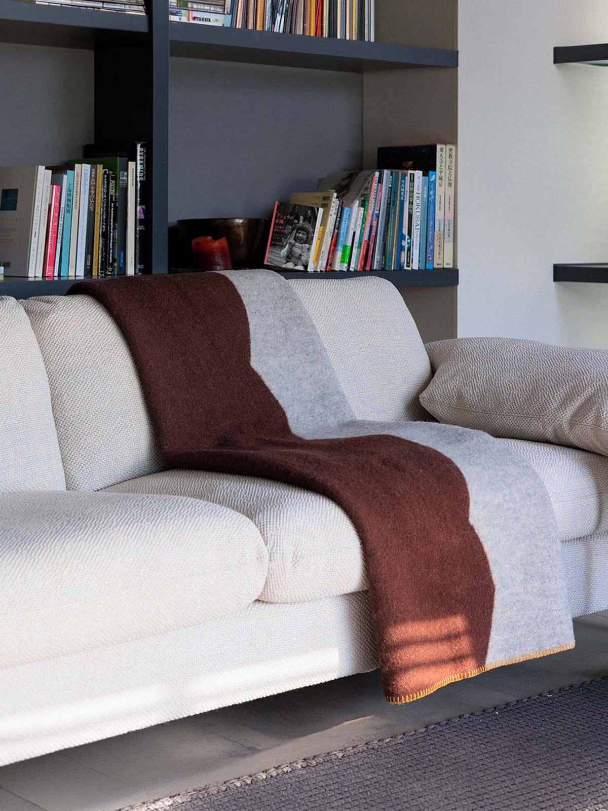 A beige sofa with a Sankt Petri Facade Wool Throw in Dark Brick draped over it sits in front of shelves filled with books and a candle.