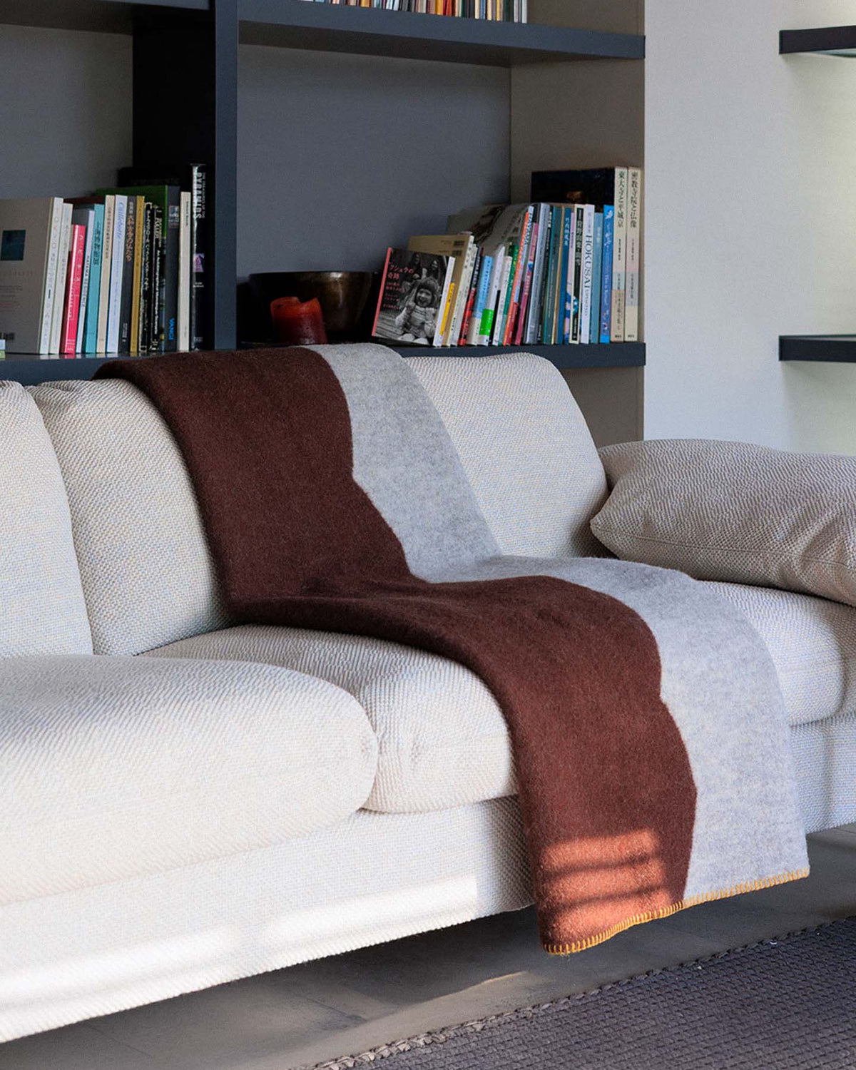 A beige sofa with a Sankt Petri Facade Wool Throw in Dark Brick draped over it sits in front of shelves filled with books and a candle.