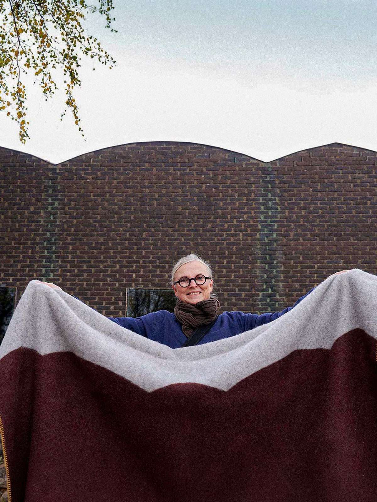 A person wearing glasses holds up a large Sankt Petri Facade Wool Throw – Dark Brick outside in front of a brown brick building.