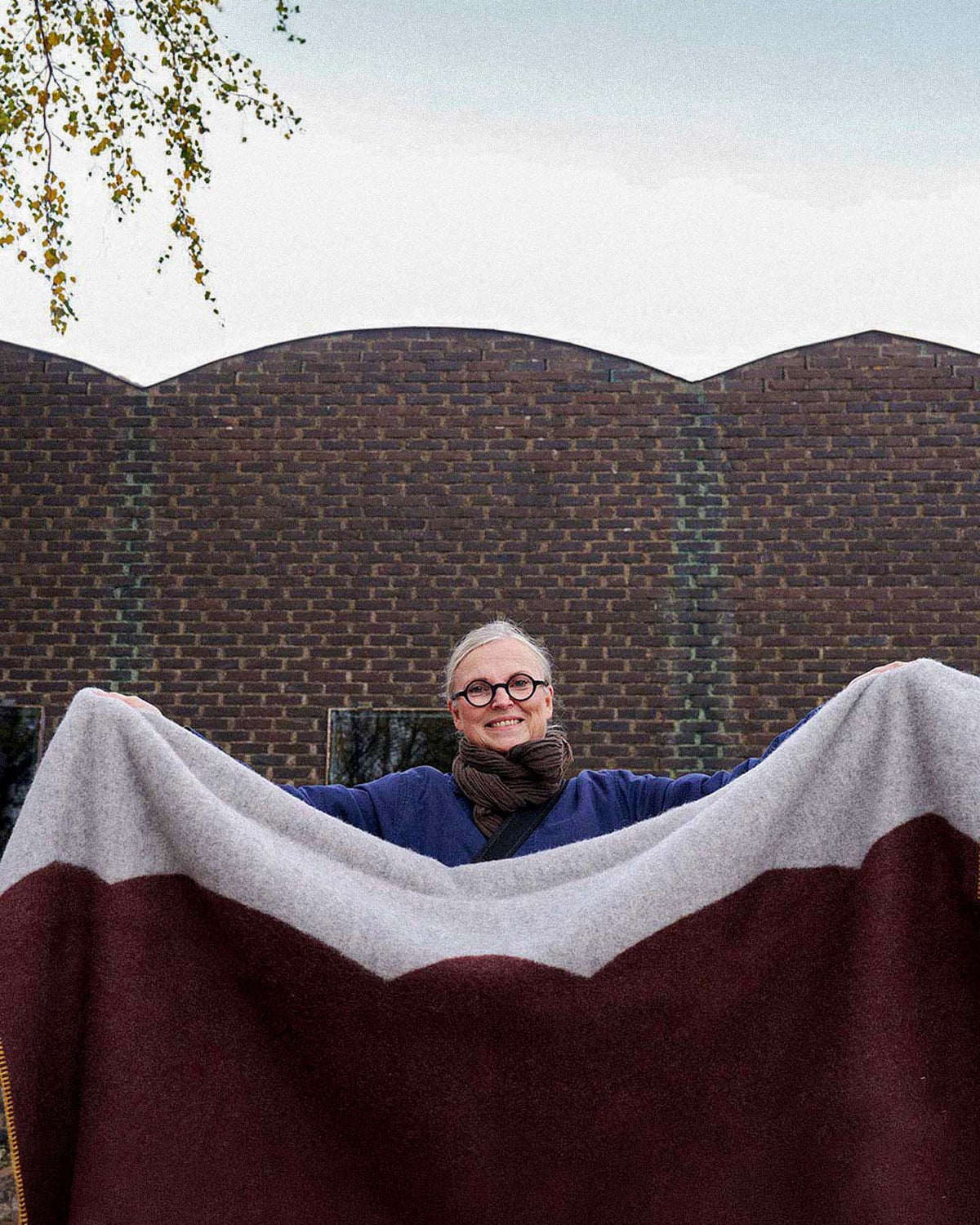 A person wearing glasses holds up a large Sankt Petri Facade Wool Throw – Dark Brick outside in front of a brown brick building.
