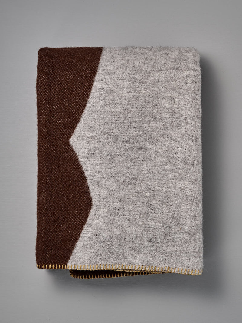 The Sankt Petri Facade Wool Throw – Dark Brick is a folded blanket crafted from organic wool, featuring a gray body accented by a brown section, highlighted with a zigzag edge and yellow stitching along the bottom—an elegant design by Kristina Stark.