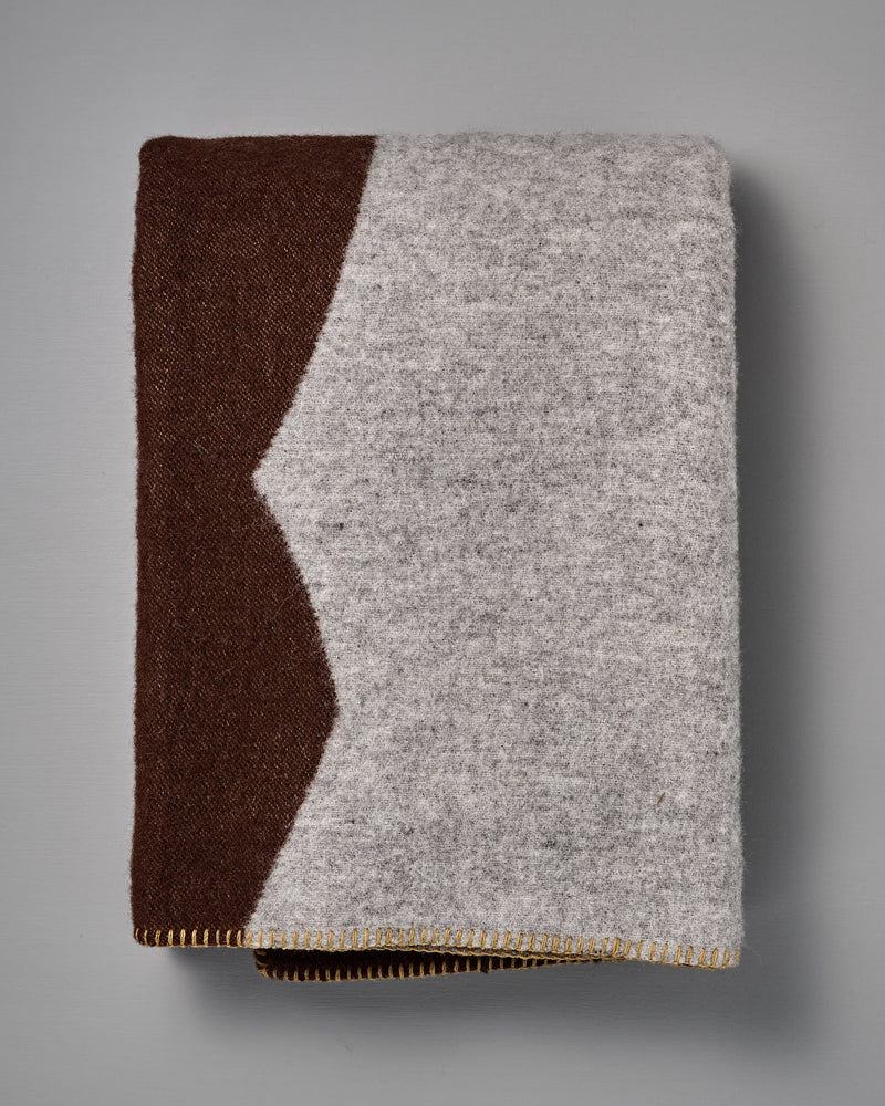 The Sankt Petri Facade Wool Throw – Dark Brick is a folded blanket crafted from organic wool, featuring a gray body accented by a brown section, highlighted with a zigzag edge and yellow stitching along the bottom—an elegant design by Kristina Stark.