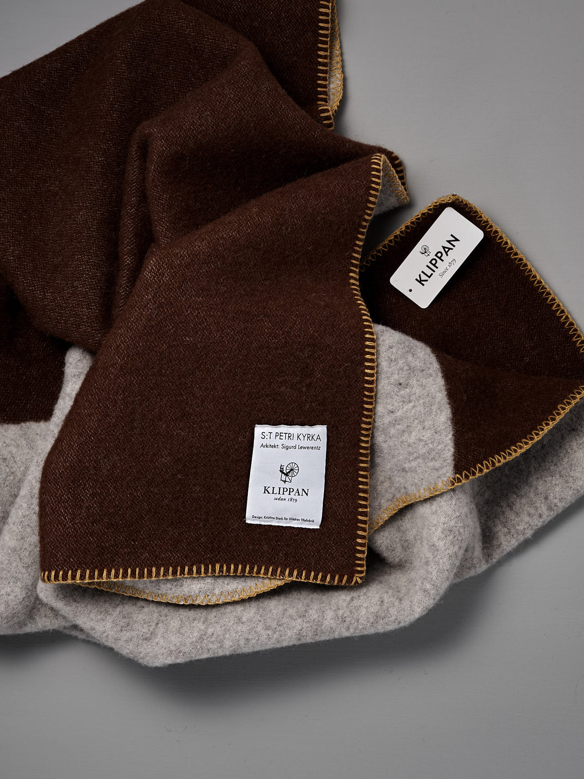 A folded Sankt Petri Facade Wool Throw – Dark Brick, made from organic wool, features brown and gray sections, yellow stitching, and two sewn labels reading “KLIPPAN” and “ST PETRI KYRKA” on a gray background—a cozy Kristina Stark design perfect for any home.