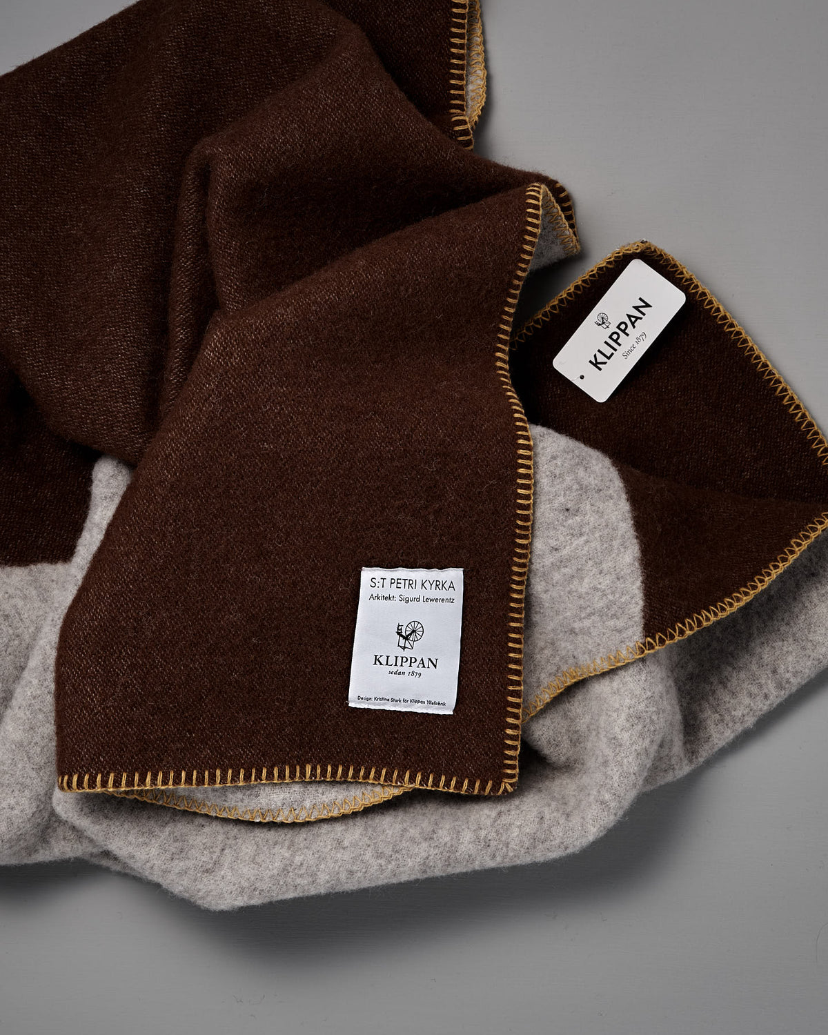 A folded Sankt Petri Facade Wool Throw – Dark Brick, made from organic wool, features brown and gray sections, yellow stitching, and two sewn labels reading “KLIPPAN” and “ST PETRI KYRKA” on a gray background—a cozy Kristina Stark design perfect for any home.