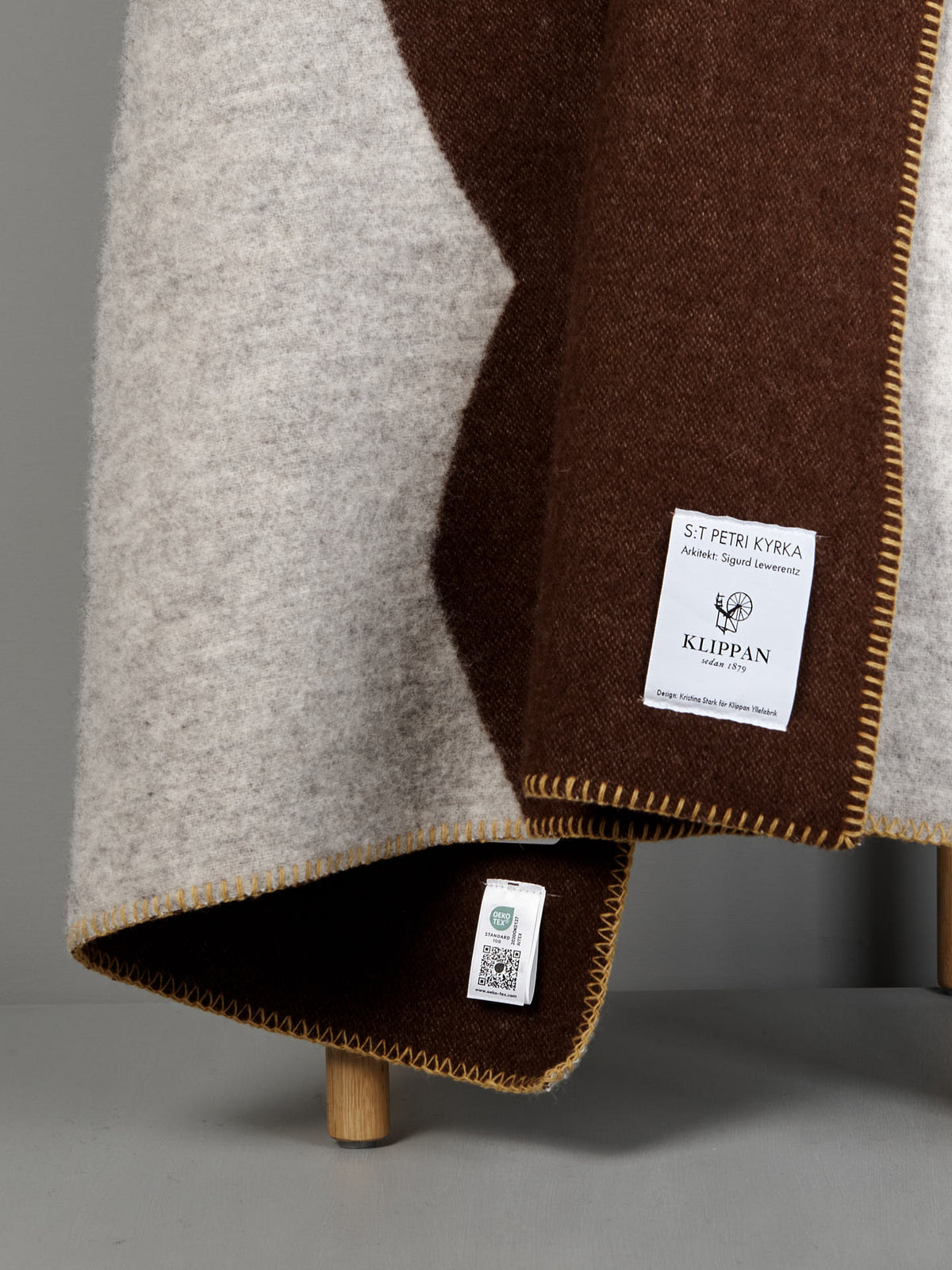 A close-up of the Sankt Petri Facade Wool Throw – Dark Brick draped over a wooden stool, showing brown and cream tones, stitched edges, and two labels attached to the organic wool fabric.