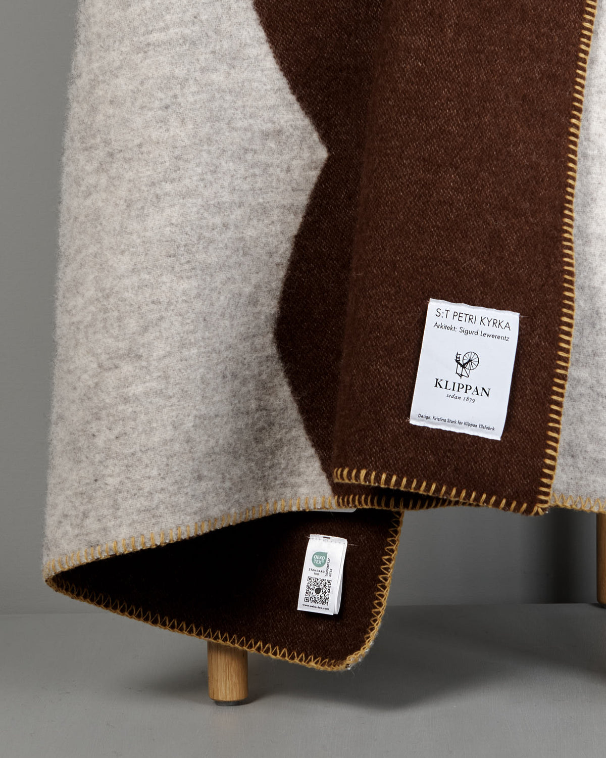 A close-up of the Sankt Petri Facade Wool Throw – Dark Brick draped over a wooden stool, showing brown and cream tones, stitched edges, and two labels attached to the organic wool fabric.