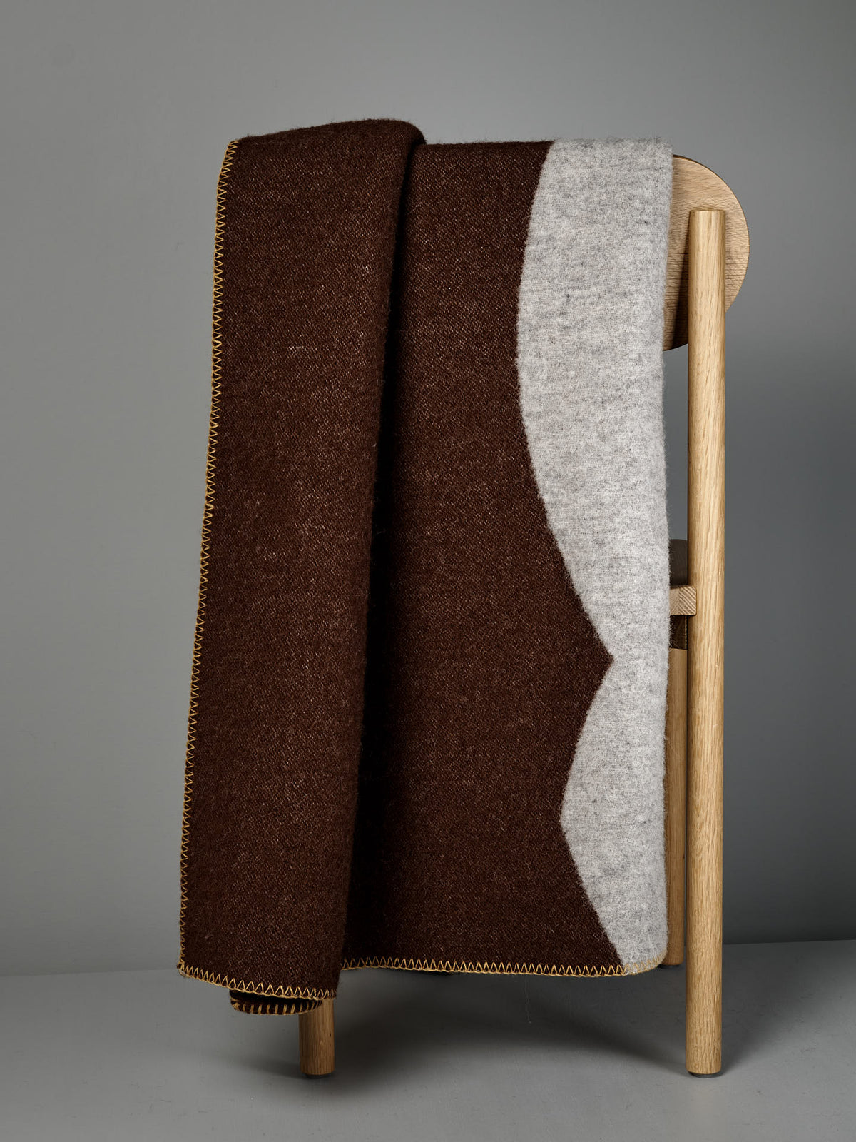 The Sankt Petri Facade Wool Throw in Dark Brick is draped over the back of a light wooden chair against a gray background.