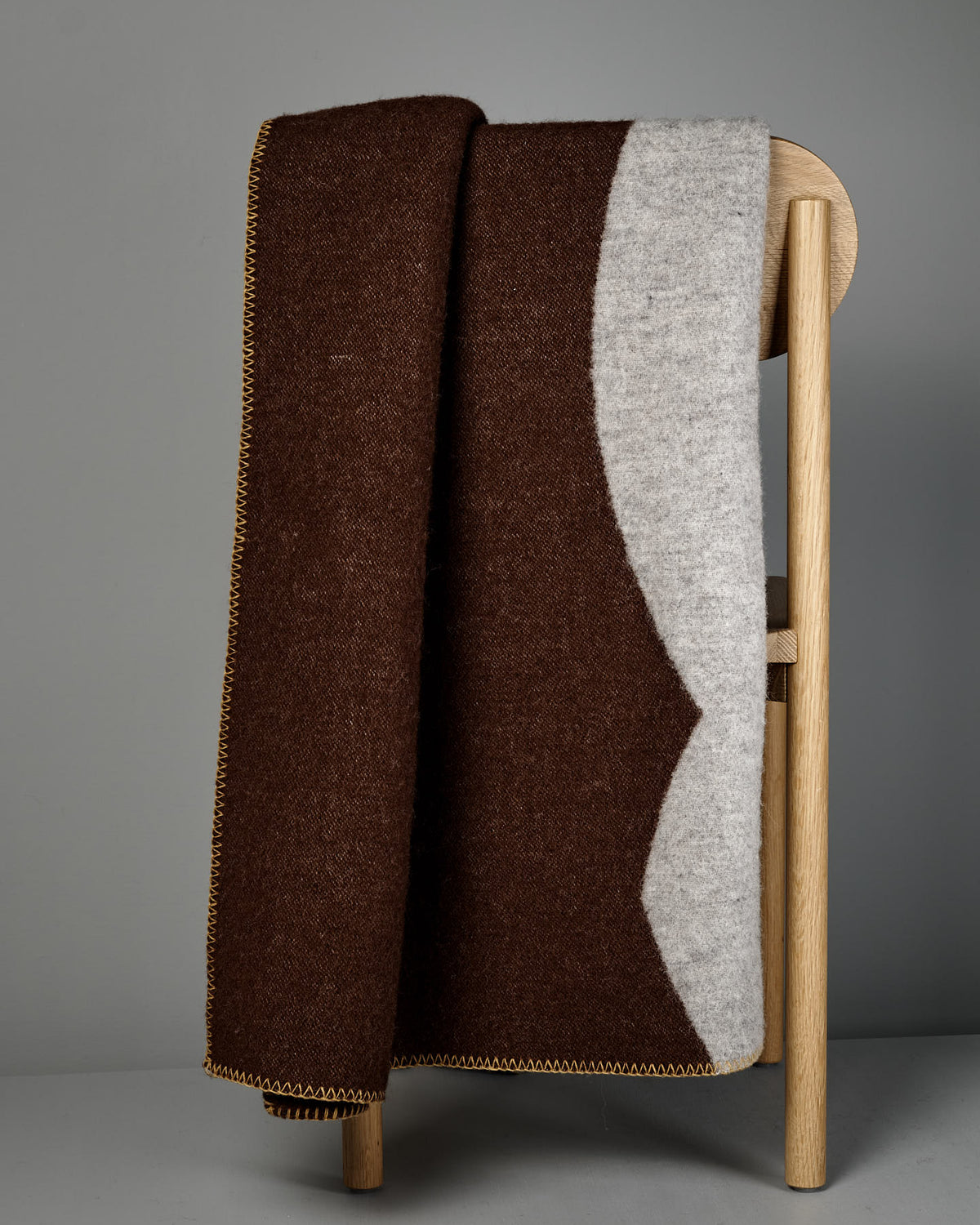 The Sankt Petri Facade Wool Throw in Dark Brick is draped over the back of a light wooden chair against a gray background.