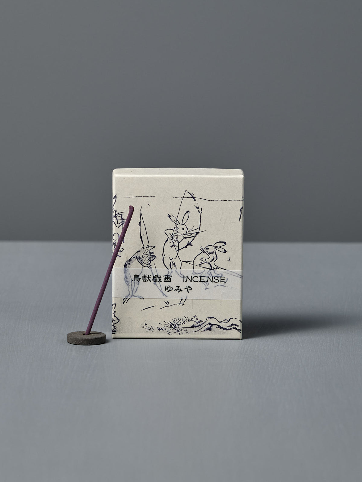 A box of Japanese Incense Yumiya – Warm Wood & Gentle Spice, featuring Japanese text and Choju Jinbutsu Giga-inspired art, stands behind a lit purple stick on a holder, releasing subtle woody fragrance against a gray backdrop.