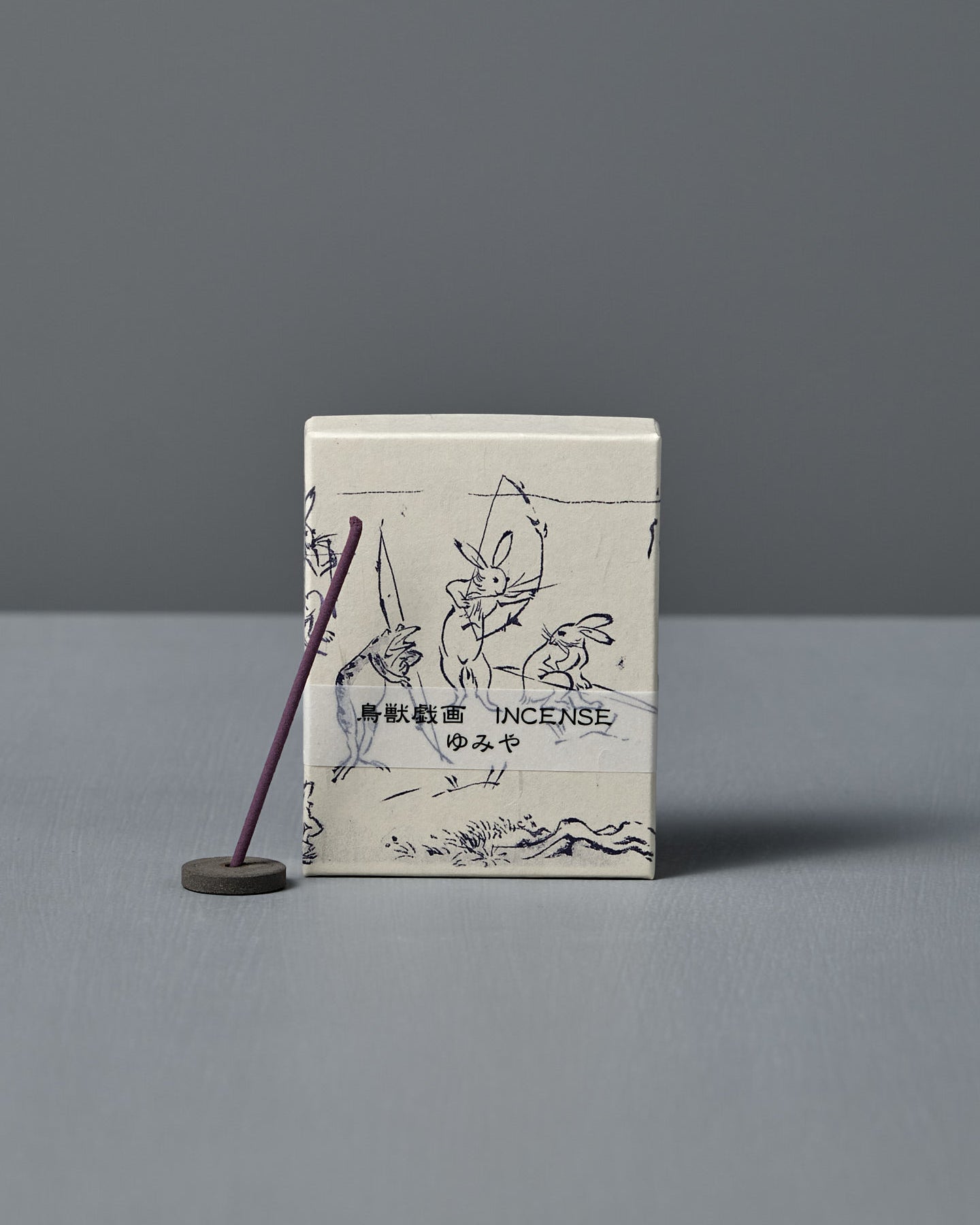 A box of Japanese Incense Yumiya – Warm Wood & Gentle Spice, featuring Japanese text and Choju Jinbutsu Giga-inspired art, stands behind a lit purple stick on a holder, releasing subtle woody fragrance against a gray backdrop.