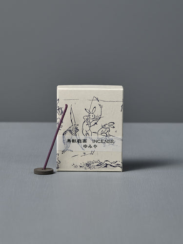 A box of Japanese Incense Yumiya – Warm Wood & Gentle Spice, featuring Japanese text and Choju Jinbutsu Giga-inspired art, stands behind a lit purple stick on a holder, releasing subtle woody fragrance against a gray backdrop.