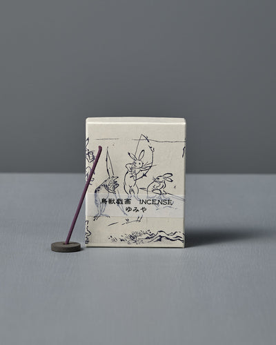 A box of Japanese Incense Yumiya – Warm Wood & Gentle Spice, featuring Japanese text and Choju Jinbutsu Giga-inspired art, stands behind a lit purple stick on a holder, releasing subtle woody fragrance against a gray backdrop.