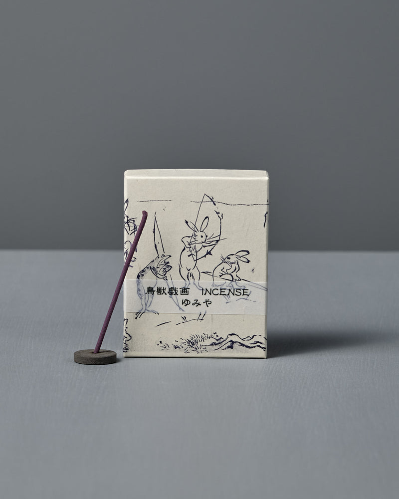 A box of Japanese Incense Yumiya – Warm Wood & Gentle Spice, featuring Japanese text and Choju Jinbutsu Giga-inspired art, stands behind a lit purple stick on a holder, releasing subtle woody fragrance against a gray backdrop.