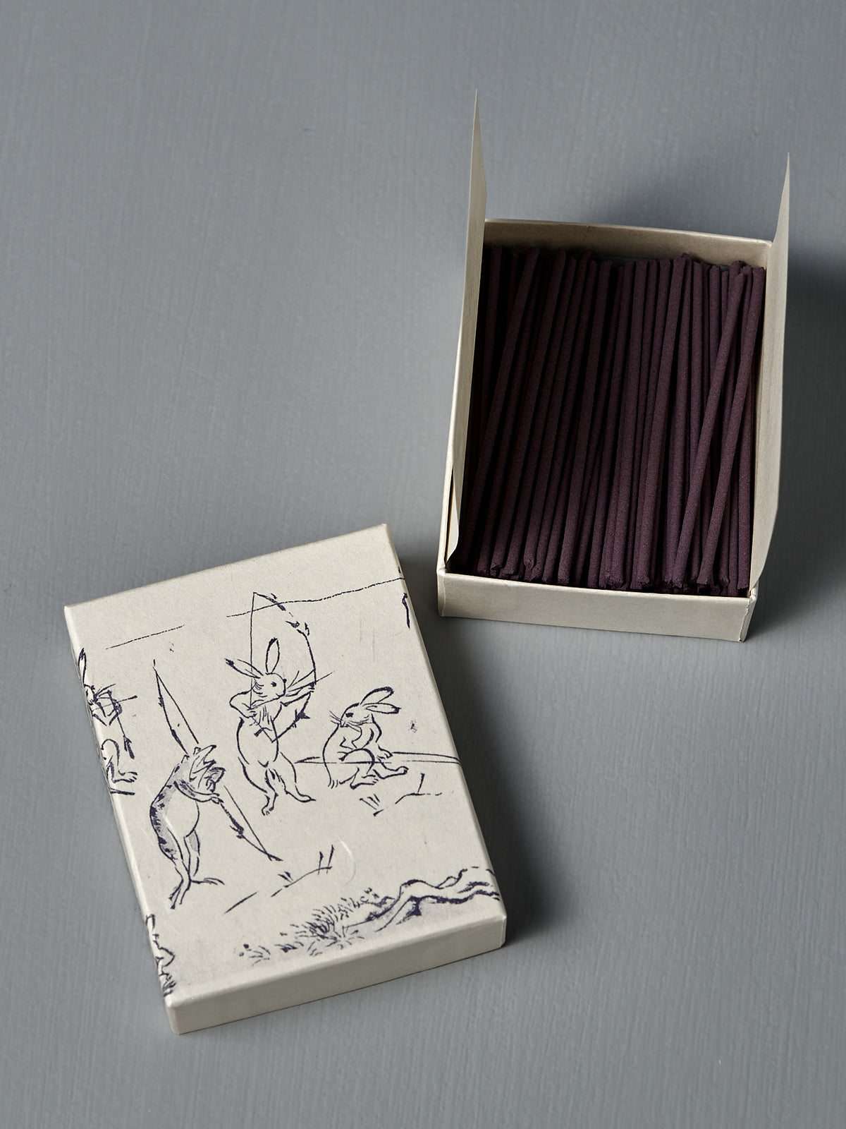 An open box of Japanese Incense Yumiya – Warm Wood & Gentle Spice sits beside a lid adorned with rabbit line art inspired by Choju Jinbutsu Giga, on a gray surface, evoking an elegant Japanese woody fragrance.