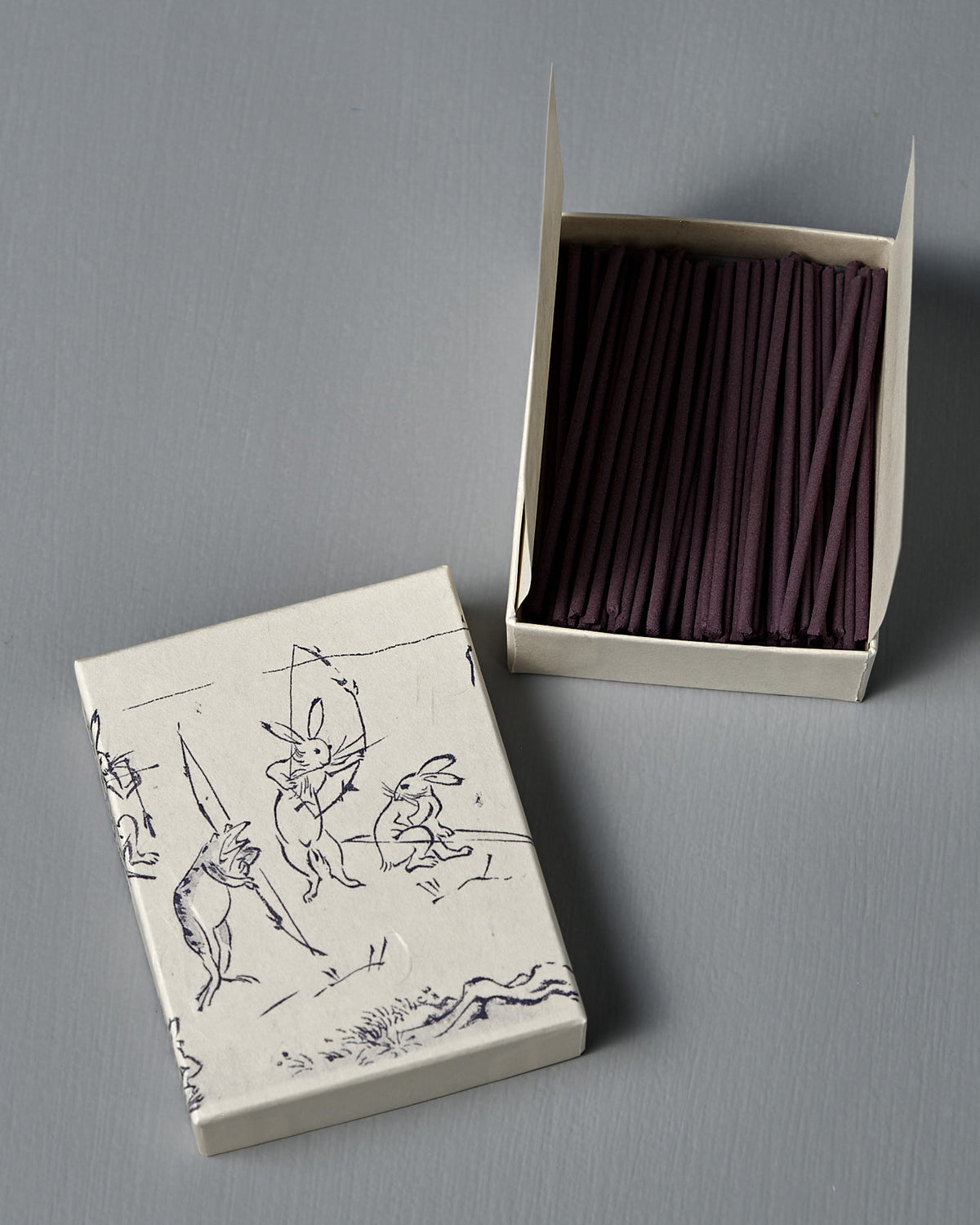 An open box of Japanese Incense Yumiya – Warm Wood & Gentle Spice sits beside a lid adorned with rabbit line art inspired by Choju Jinbutsu Giga, on a gray surface, evoking an elegant Japanese woody fragrance.