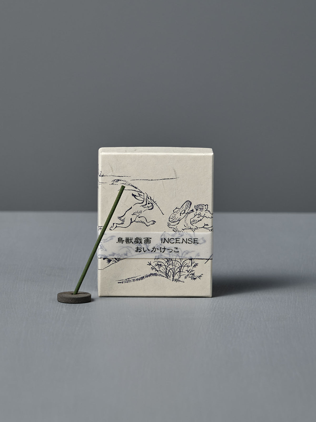 A box of Japanese Incense Oikakekko – Fresh Green & Soft Wood with minimalist designs and Japanese text stands upright behind a lit incense stick in a holder on a gray surface, adding a subtle green herbal scent.