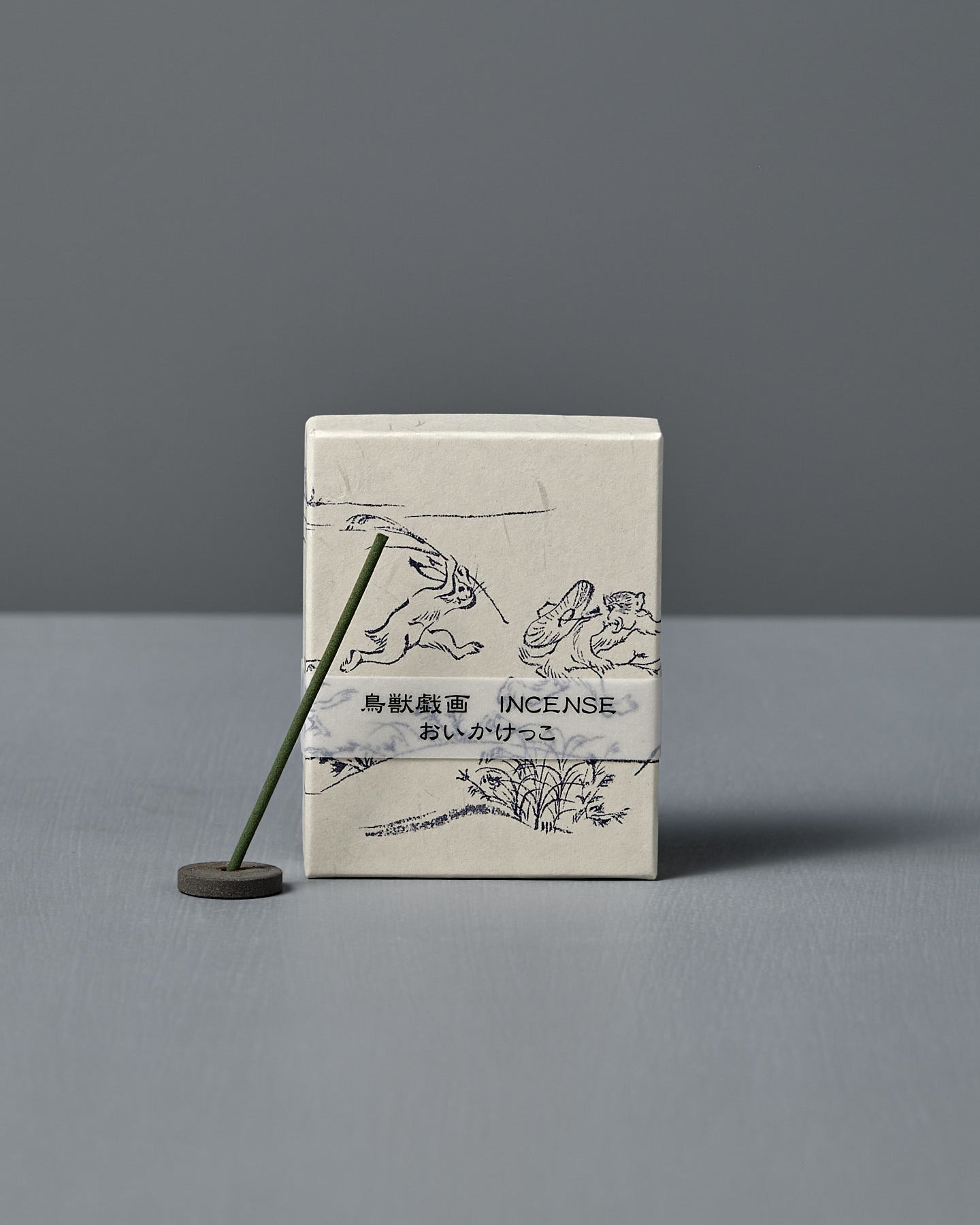A box of Japanese Incense Oikakekko – Fresh Green & Soft Wood with minimalist designs and Japanese text stands upright behind a lit incense stick in a holder on a gray surface, adding a subtle green herbal scent.