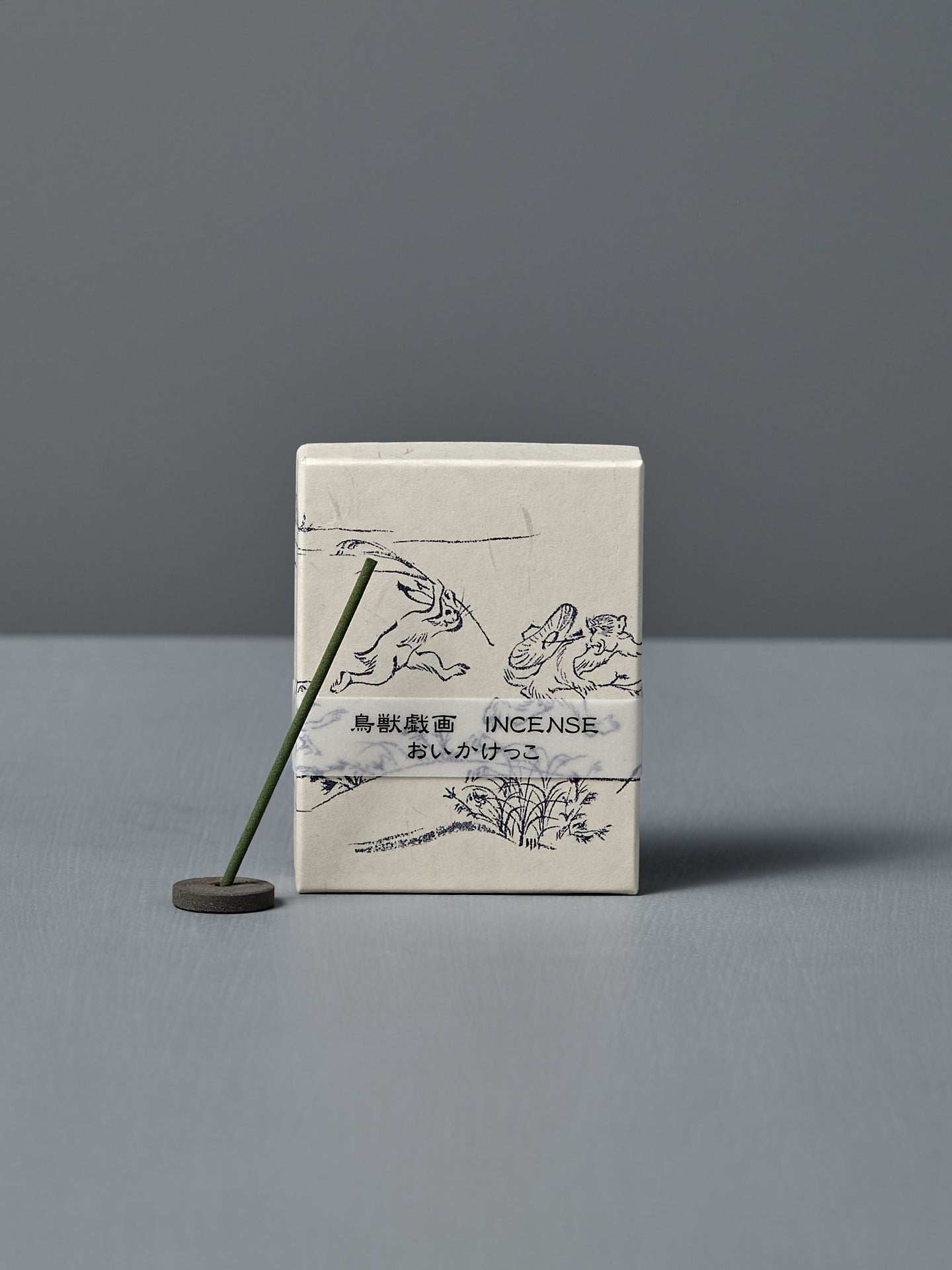 A box of Japanese Incense Oikakekko – Fresh Green & Soft Wood with minimalist designs and Japanese text stands upright behind a lit incense stick in a holder on a gray surface, adding a subtle green herbal scent.