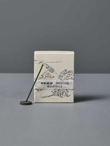 A box of Japanese Incense Oikakekko – Fresh Green & Soft Wood with minimalist designs and Japanese text stands upright behind a lit incense stick in a holder on a gray surface, adding a subtle green herbal scent.