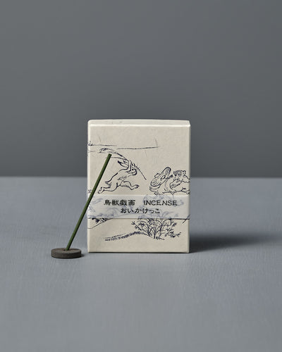 A box of Japanese Incense Oikakekko – Fresh Green & Soft Wood with minimalist designs and Japanese text stands upright behind a lit incense stick in a holder on a gray surface, adding a subtle green herbal scent.