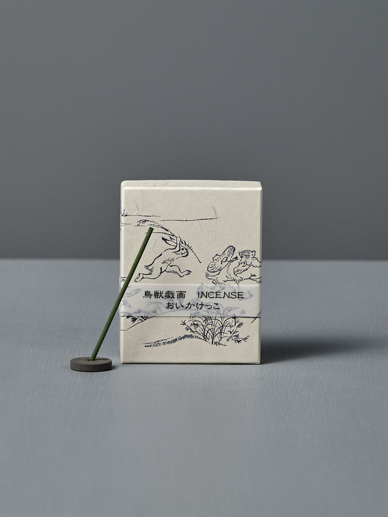 A box of Japanese Incense Oikakekko – Fresh Green & Soft Wood with minimalist designs and Japanese text stands upright behind a lit incense stick in a holder on a gray surface, adding a subtle green herbal scent.