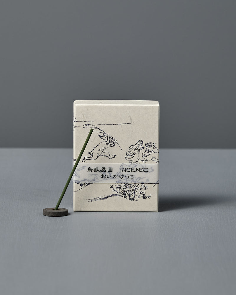 A box of Japanese Incense Oikakekko – Fresh Green & Soft Wood with minimalist designs and Japanese text stands upright behind a lit incense stick in a holder on a gray surface, adding a subtle green herbal scent.