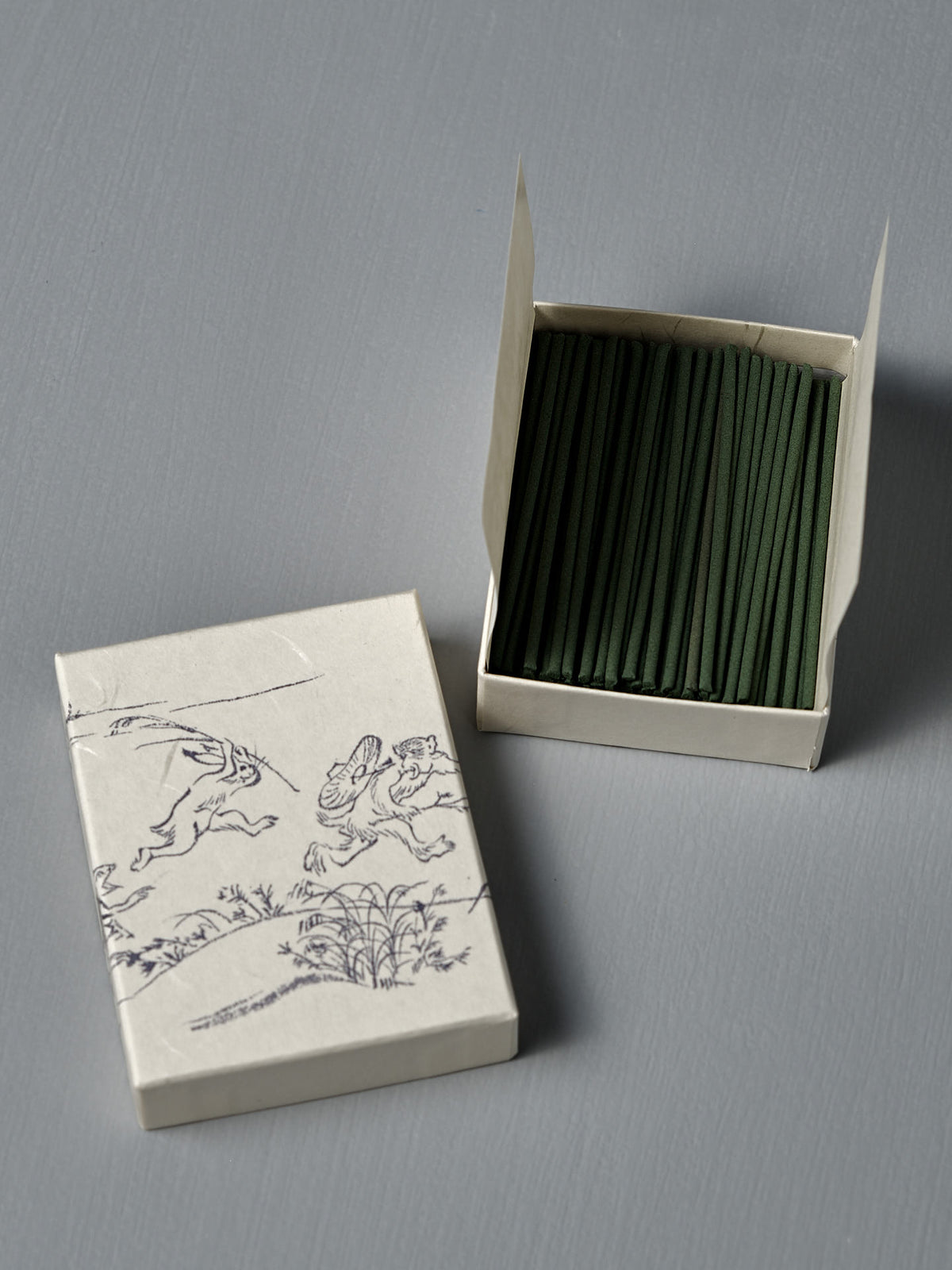 An open box of Japanese Incense Oikakekko – Fresh Green & Soft Wood sits beside its lid, which displays simple line art of animals and plants—classic Japanese incense with a vibrant green herbal and soft wood aroma.