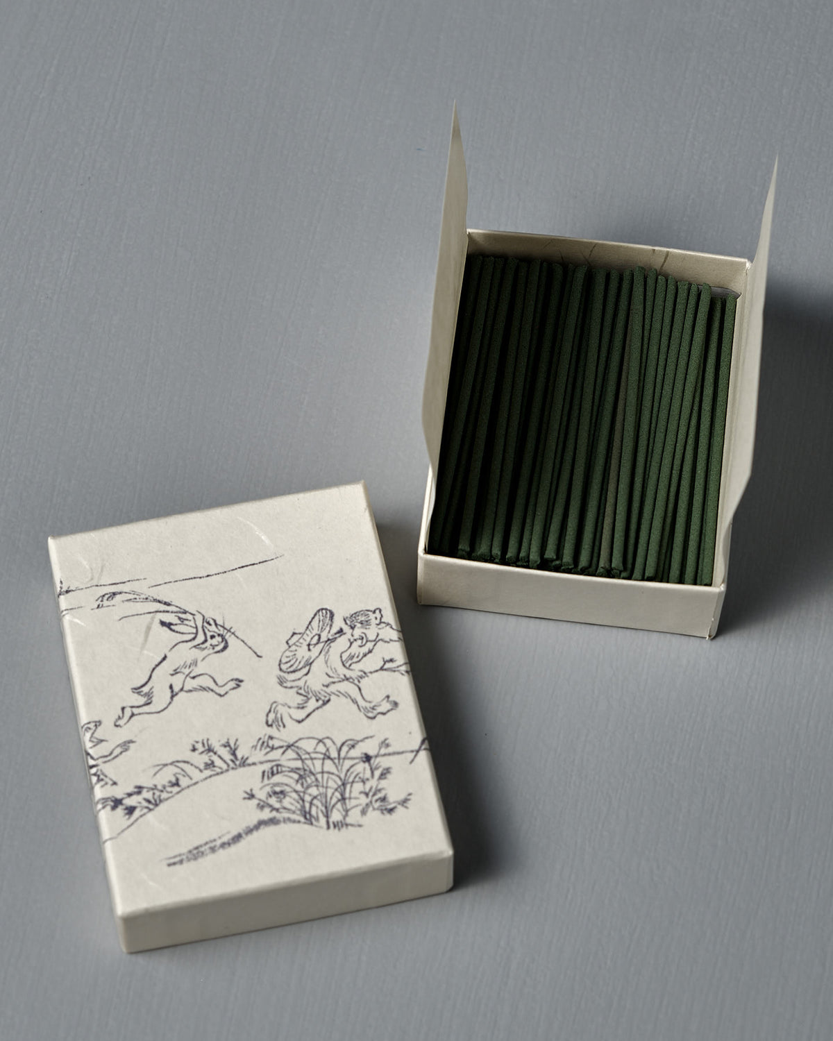 An open box of Japanese Incense Oikakekko – Fresh Green & Soft Wood sits beside its lid, which displays simple line art of animals and plants—classic Japanese incense with a vibrant green herbal and soft wood aroma.