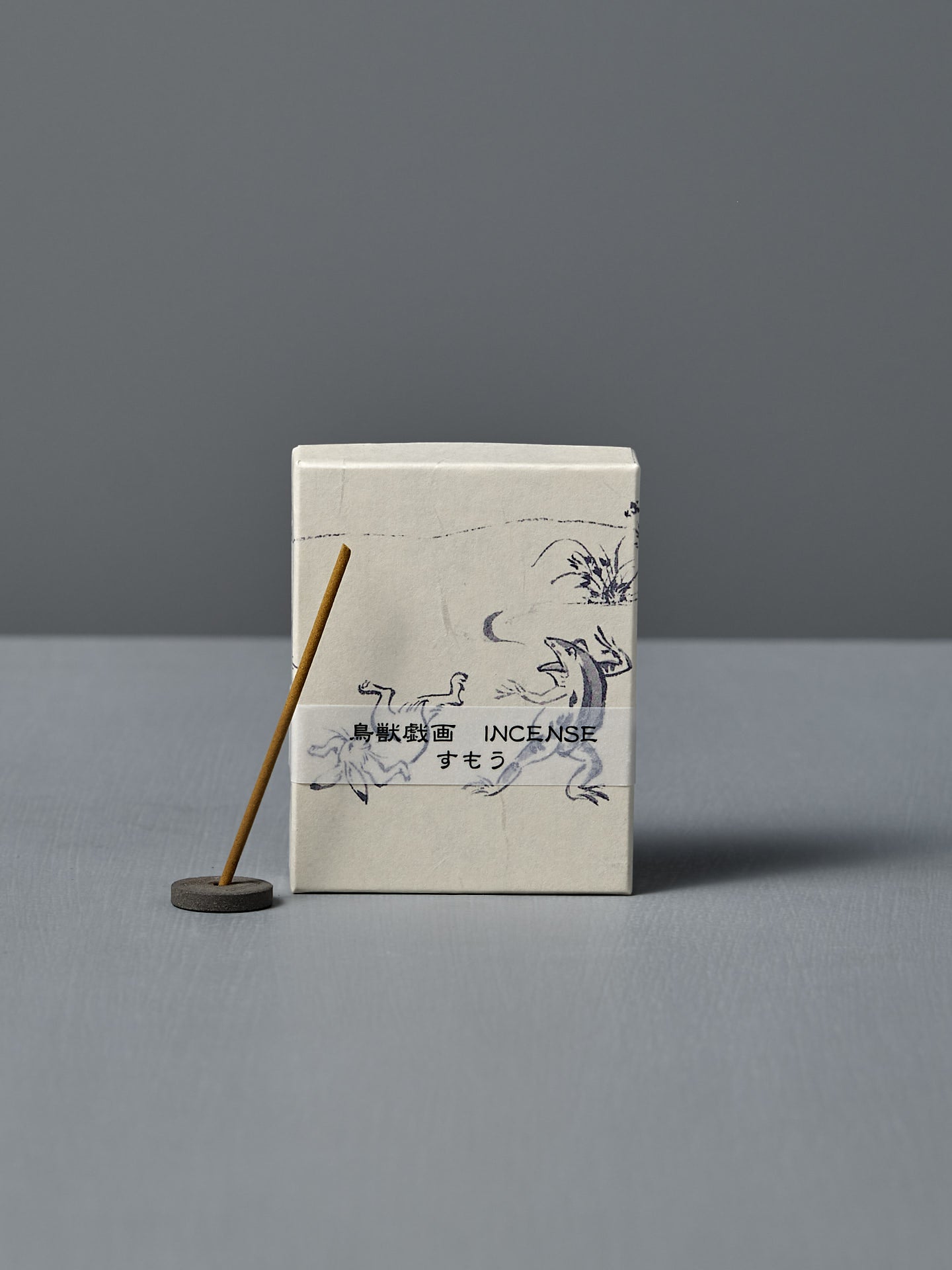 A box of Japanese Incense Sumo – Pear, Calone & Sandalwood, featuring Japanese text and animal illustrations, sits beside a lit incense stick in a holder on a gray surface, releasing a subtle aquatic-fruity aroma.