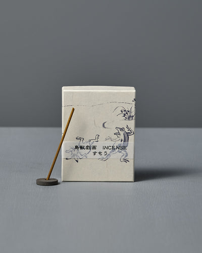A box of Japanese Incense Sumo – Pear, Calone & Sandalwood, featuring Japanese text and animal illustrations, sits beside a lit incense stick in a holder on a gray surface, releasing a subtle aquatic-fruity aroma.