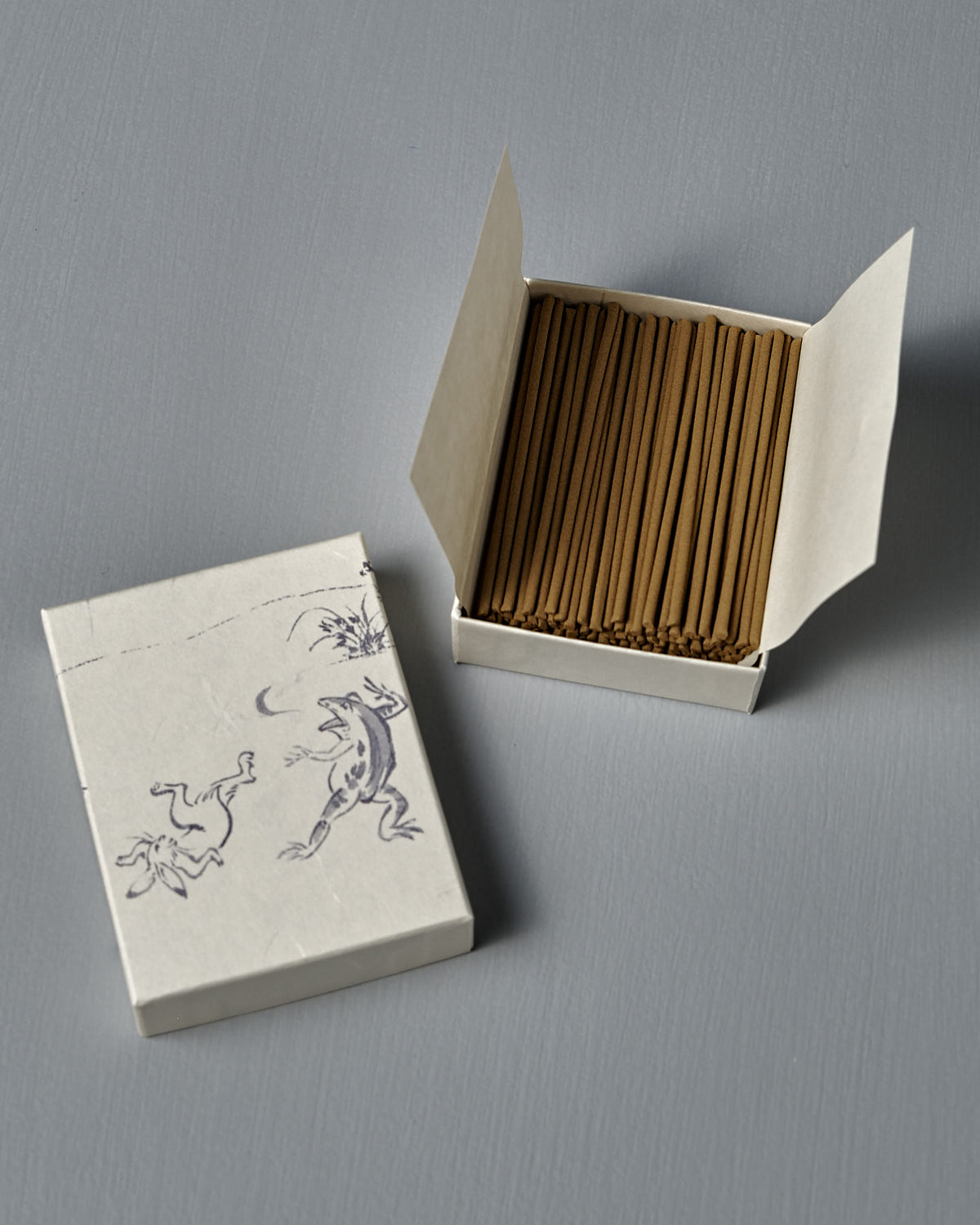 Open box of Japanese Incense Sumo – Pear, Calone & Sandalwood, featuring two frogs illustrated on the lid and placed on a gray surface; sandalwood aroma combines with fresh pear and aquatic notes.