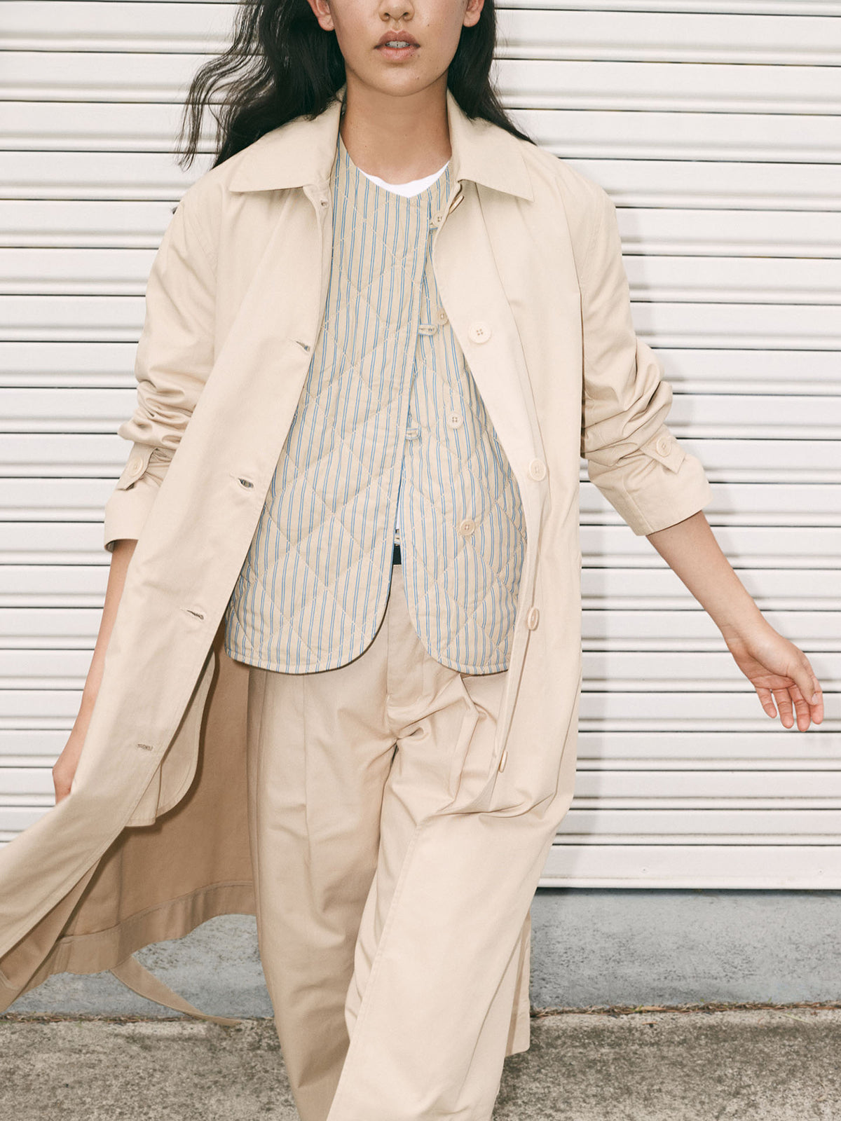 A person wears the Kowtow Axis Vest – Double Pinstripe over a striped shirt and beige pants, standing before a white metal shutter.
