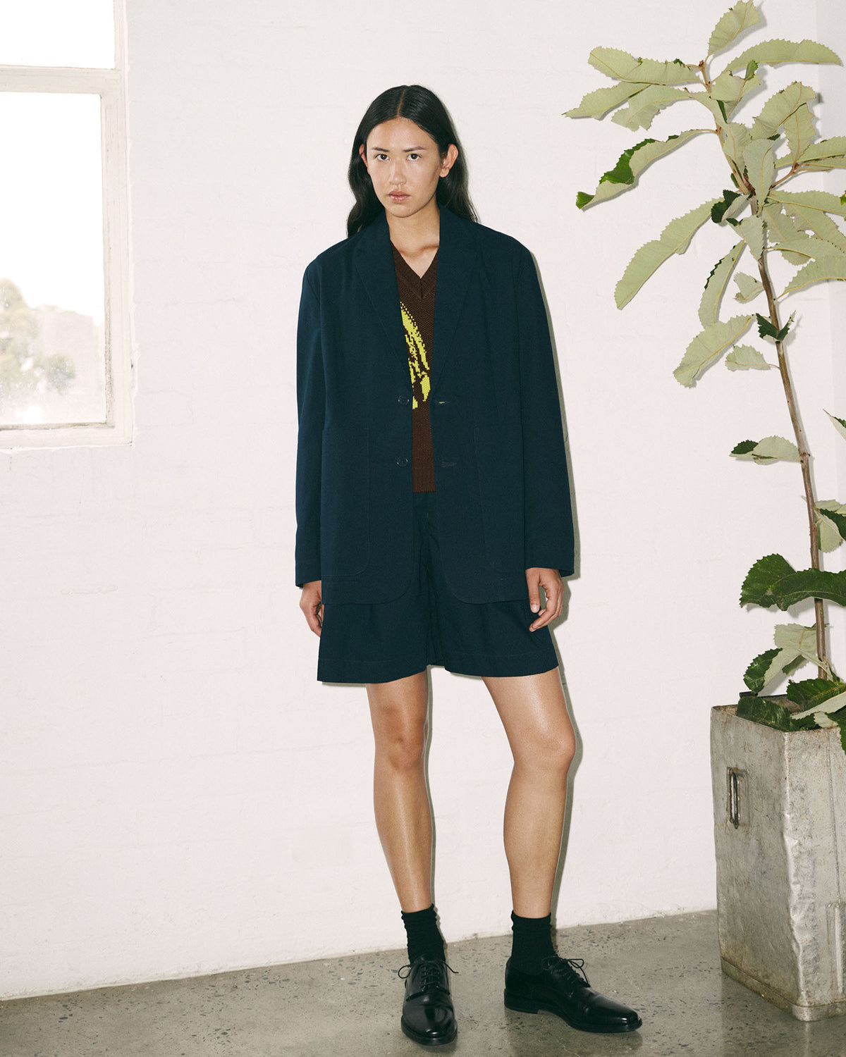 A person stands indoors wearing the Kowtow Carter Blazer in Evening Navy, styled with shorts, a brown oversized sweater with yellow detail, black socks, and black shoes; a potted plant and window are in the background.