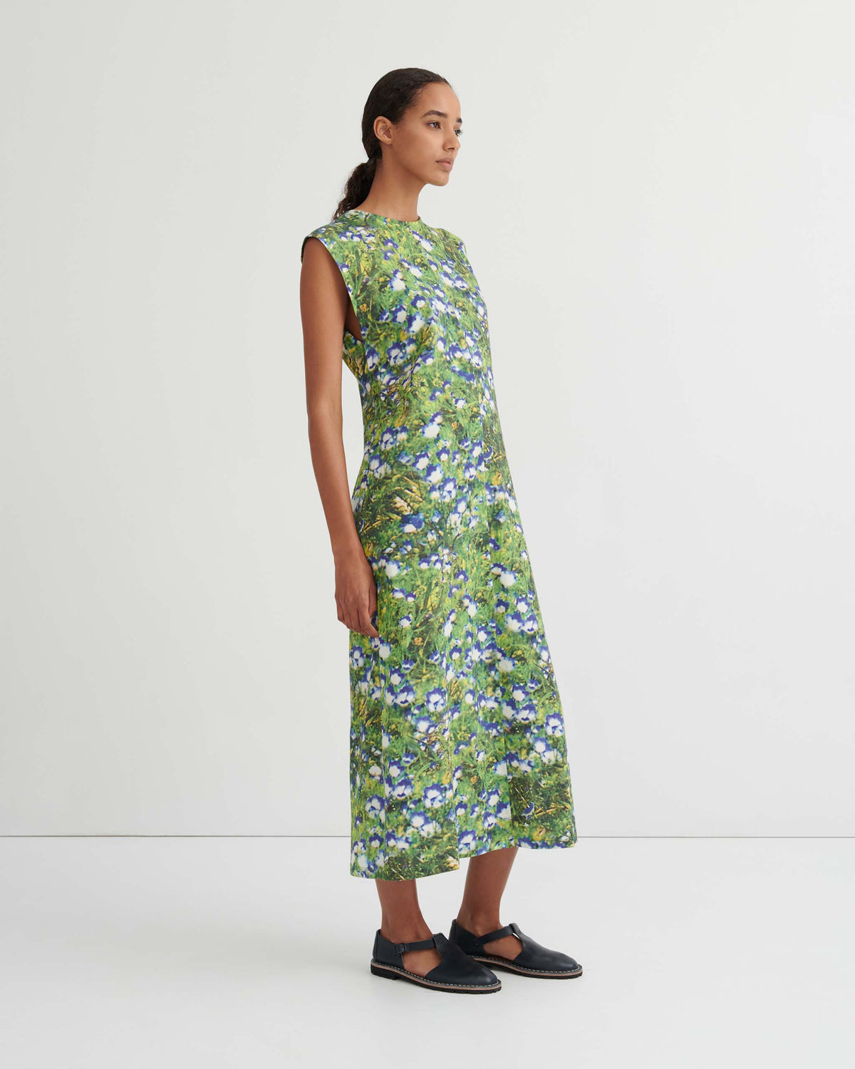 Woman standing in a sleeveless, calf-length dress with a green and blue floral pattern, black shoes, and hair pulled back, against a plain white background.
