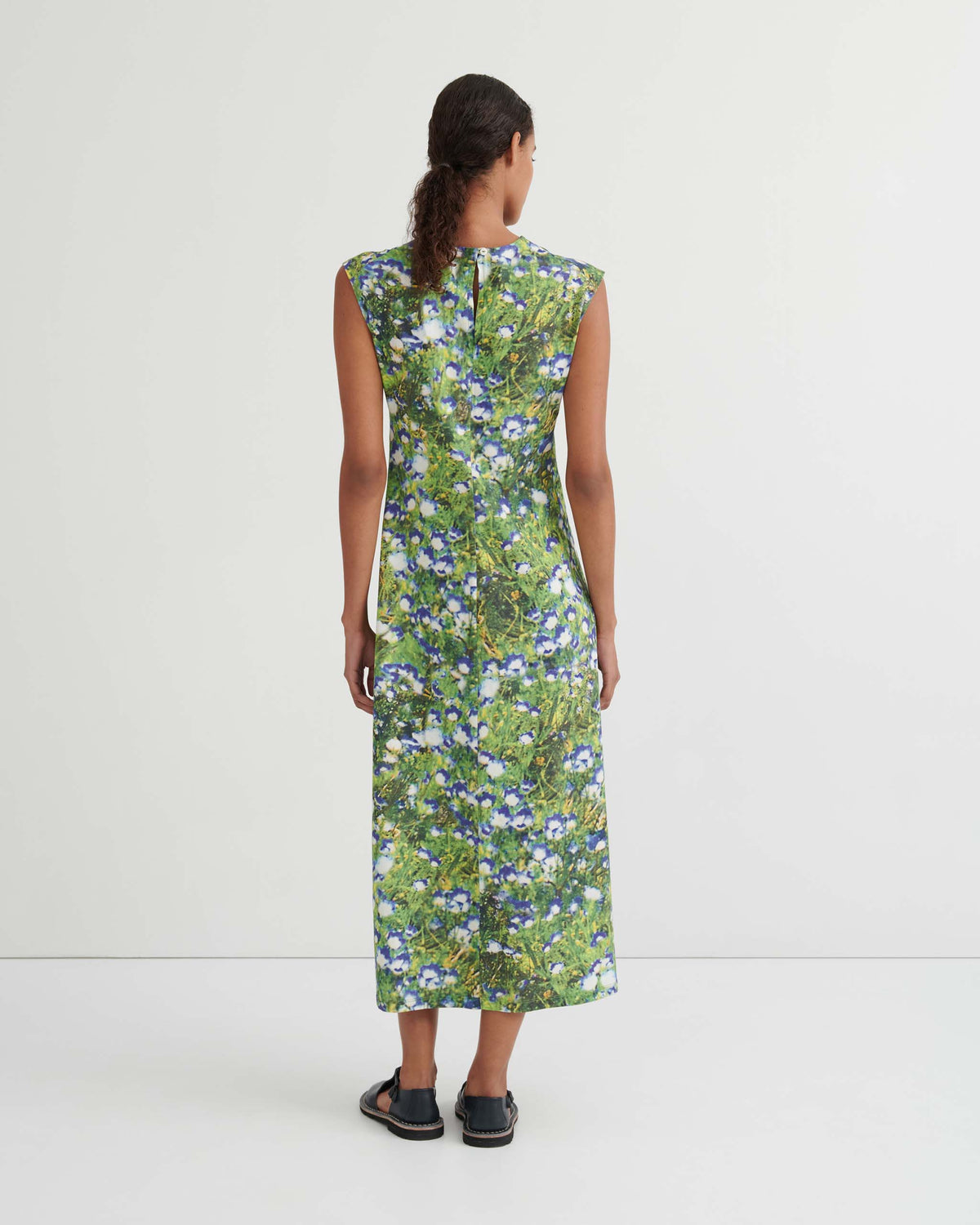 Person standing against a white wall, shown from the back, wearing a sleeveless, ankle-length dress with a green and blue floral pattern and black sandals.