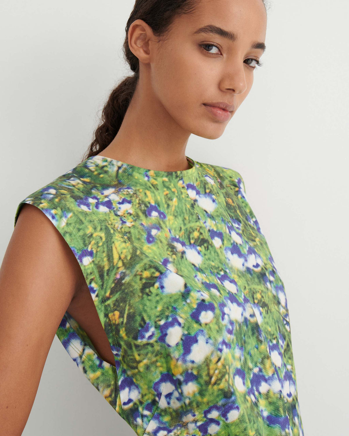 Woman wearing a sleeveless top with a green and blue floral print, standing against a plain white background, looking at the camera.