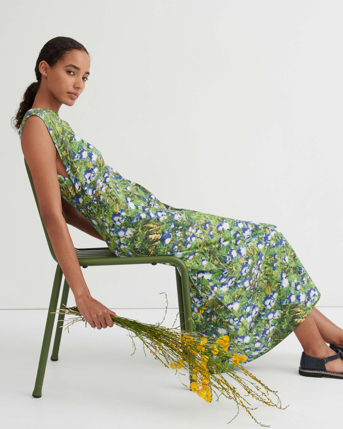 Woman in a green floral dress sits sideways on a green chair, holding a bunch of yellow flowers, against a plain white background.