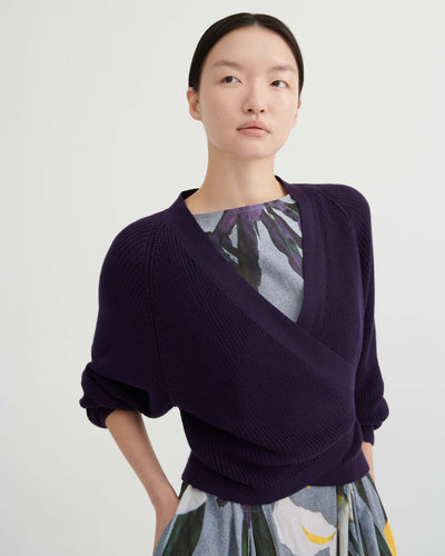 A person poses against a plain background wearing the Composure Cardigan – Dahlia by Kowtow in a relaxed fit, layered over a floral-patterned dress with a gray base and yellow accents.