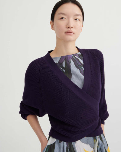 A person poses against a plain background wearing the Composure Cardigan – Dahlia by Kowtow in a relaxed fit, layered over a floral-patterned dress with a gray base and yellow accents.