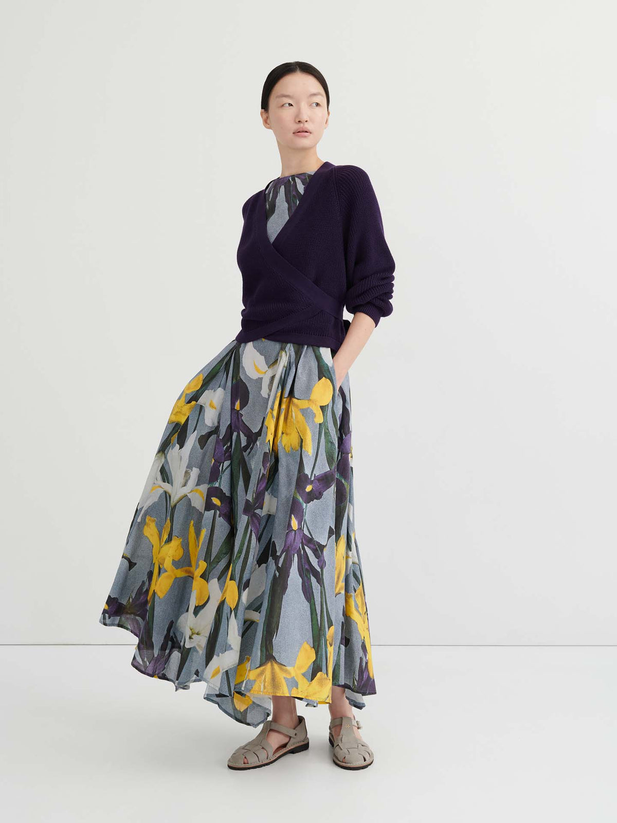 A woman wearing a relaxed fit dark purple Composure Cardigan – Dahlia over a long, flowy floral dress with yellow and purple flowers, paired with beige sandals, stands against a plain white background.