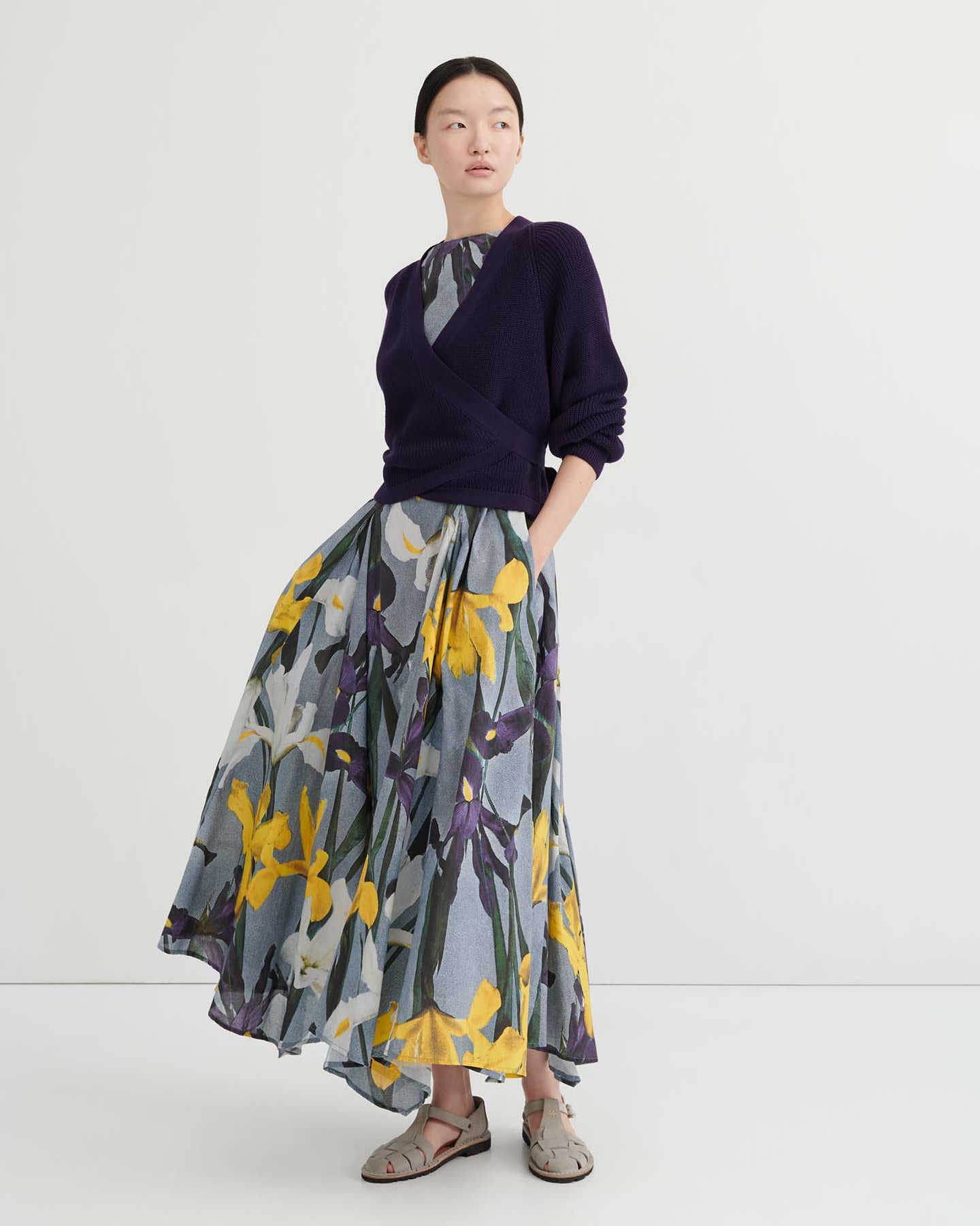 A person poses against a plain background wearing the Composure Cardigan – Dahlia by Kowtow in a relaxed fit, layered over a floral-patterned dress with a gray base and yellow accents.