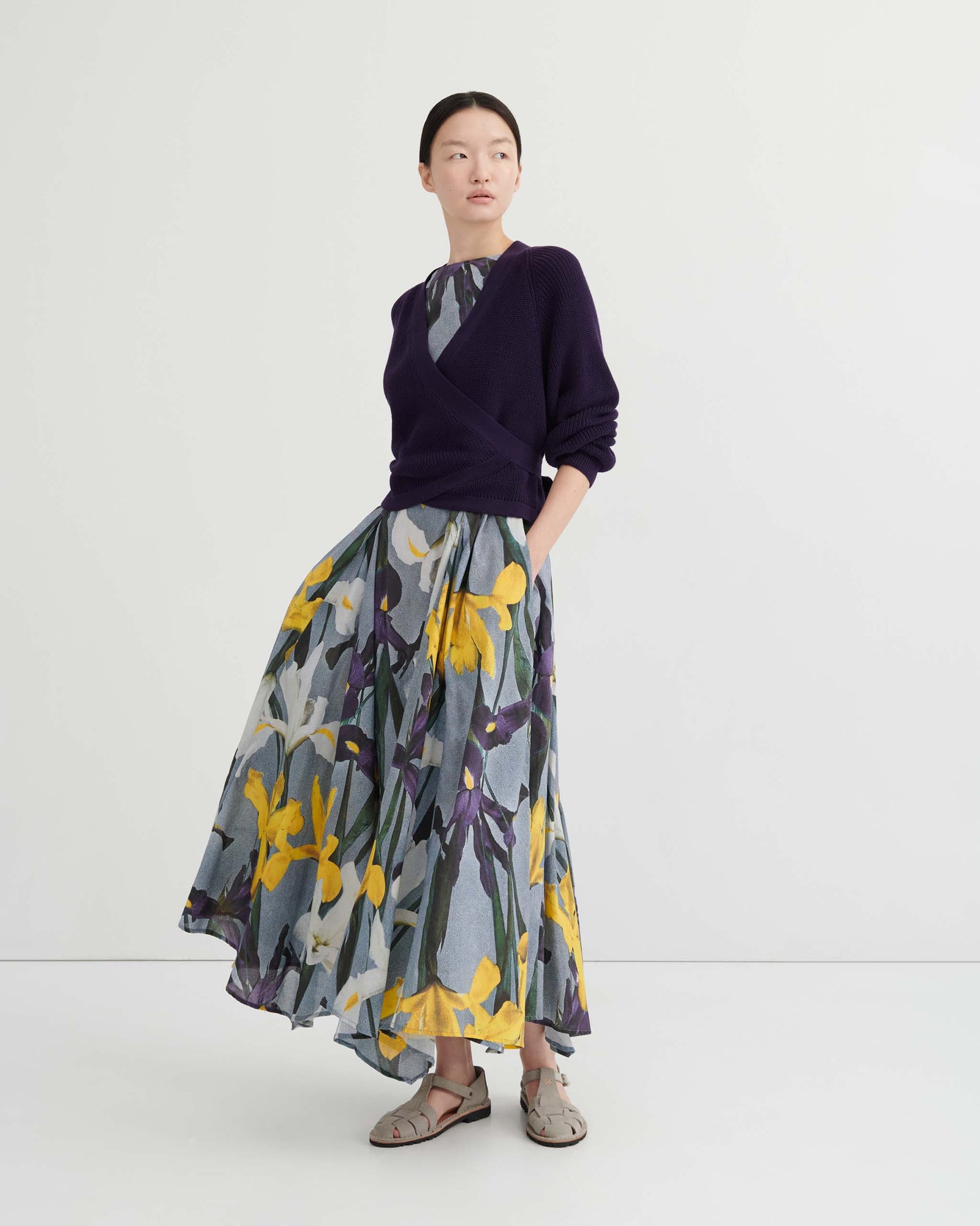 A person poses against a plain background wearing the Composure Cardigan – Dahlia by Kowtow in a relaxed fit, layered over a floral-patterned dress with a gray base and yellow accents.